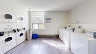Located at 10 Highland Crescent in Kitchener, Ontario, this communal laundry facility offers a practical and welcoming environment designed to serve residents efficiently. The single-floor space is thoughtfully divided into distinct functional zones, all unified by light wood-style flooring, bright white walls, and a clean, modern aesthetic throughout.

The main laundry area is well-equipped with a full row of front-loading washing machines and dryers, including stacked units that make efficient use of vertical space. A utility sink provides added convenience, while multiple stainless steel folding tables offer ample surface area for sorting and organizing laundry. Bright fluorescent lighting ensures the workspace is well-illuminated at all times.

Natural light filters through a large window in the washing area, lending a pleasant, airy feel to what might otherwise be a purely utilitarian space. Waste and recycling bins are conveniently positioned throughout.

A separate waiting and common area provides a comfortable place to relax, furnished with modern seating, round tables, and chairs in a contemporary Scandinavian style. Ceiling-mounted light fixtures cast a warm glow over the space, which is further enlivened by decorative artwork and a potted plant. Additional seating near the entrance corridors and a payment or information station complete this thoughtfully designed, resident-focused facility.
