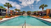 Located at 7601 N Ninth Ave in Pensacola, Florida, this outdoor pool area serves as a central recreational amenity for the surrounding residential community. The generously sized rectangular swimming pool features clear turquoise water and is accessible via built-in entry steps and a stainless steel handrail, with depth markers visible along the pool edge. The expansive concrete sun deck surrounds the pool on all sides, providing ample space for relaxation and outdoor recreation. Vibrant orange lounge chairs are arranged around the perimeter, complemented by shaded seating areas with patio umbrellas. The pool area is fully enclosed by a black iron fence, ensuring security and defined boundaries. A utility and storage structure is positioned centrally along one side, alongside posted pool rules signage. Towering palm trees and mature shade trees line the fencing, adding a lush, tropical character fitting of the Florida Gulf Coast climate. The surrounding two-story residential buildings frame the space on multiple sides, creating an intimate courtyard-like atmosphere. The area benefits from abundant natural sunlight throughout the day, making it an inviting outdoor destination. The overall setting combines functional amenities with a relaxed, resort-inspired aesthetic well-suited to the warm Pensacola climate.