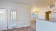 This single-story residence offers a practical and comfortable layout with a clean, neutral aesthetic throughout. The living area is a standout space, featuring soaring vaulted ceilings, soft carpet flooring, and a ceiling fan overhead. A large window fills the room with natural light, while a glass-paneled exterior door provides additional brightness and outdoor access. The open flow connects the living space to the dining area, which is elevated by a chandelier and transitions into tile flooring. A breakfast bar separates the dining space from the galley-style kitchen, which is equipped with white cabinetry, a full appliance suite including a refrigerator and range, a double sink with ample counter space, and stone-look tile flooring that extends throughout the kitchen and wet areas. The bedrooms are carpeted and well-proportioned, each with access to natural light through windows that frame views of surrounding greenery. The full bathroom features a combination tub and shower with grab bars, a single-sink vanity with storage below, and matching stone-look tile flooring. Textured ceilings and white panel doors are consistent throughout, contributing to the space's cohesive and tidy character.