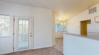 This single-story residence offers a practical and comfortable layout with a clean, neutral aesthetic throughout. The living area is a standout space, featuring soaring vaulted ceilings, soft carpet flooring, and a ceiling fan overhead. A large window fills the room with natural light, while a glass-paneled exterior door provides additional brightness and outdoor access. The open flow connects the living space to the dining area, which is elevated by a chandelier and transitions into tile flooring. A breakfast bar separates the dining space from the galley-style kitchen, which is equipped with white cabinetry, a full appliance suite including a refrigerator and range, a double sink with ample counter space, and stone-look tile flooring that extends throughout the kitchen and wet areas. The bedrooms are carpeted and well-proportioned, each with access to natural light through windows that frame views of surrounding greenery. The full bathroom features a combination tub and shower with grab bars, a single-sink vanity with storage below, and matching stone-look tile flooring. Textured ceilings and white panel doors are consistent throughout, contributing to the space's cohesive and tidy character.
