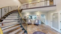 Villa Del Rio Luxury Living at 303 Greenwich Ave in Warwick, RI offers an expansive two-story amenity facility thoughtfully designed to support an active and social lifestyle. The welcoming entrance lobby greets residents with soaring ceilings, large transom windows, stone-look tile flooring, and an ornate wrought-iron staircase connecting both levels, setting an elegant tone throughout.

The first floor houses a bright lounge and café-style seating area with large windows overlooking lush greenery, a wall-mounted TV, and a mix of bistro tables and lounge chairs. A professionally appointed fitness center features an extensive array of cardio machines and strength equipment on durable rubber flooring beneath a drop-panel ceiling with ample lighting. Three well-maintained bathrooms with stone tile floors, backlit mirrors, and modern fixtures serve both levels.

The upper floor expands the social offerings with multiple living rooms, including a spacious bar lounge featuring hardwood floors, a long granite-topped bar with pendant lighting, and plush velvet seating. A cozy lounge area offers sectional seating and colorful wall art. The game room stands out with a full-size pool table, a rustic reclaimed-wood accent wall, flat-screen TV, and bar-height seating. A private office with hardwood floors and garden views rounds out the upper level. Two hallways provide seamless circulation throughout.
