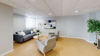 Located at 1145-1175 Adair Ct in Windsor, Ontario, this single-floor space offers a thoughtfully organized layout designed for functional communal use. The interior is finished with light hardwood-style flooring throughout, neutral grey walls, and a drop tile ceiling with integrated recessed lighting that provides consistent, even illumination across the entire space.

The open main area serves as a versatile lounge, furnished with a sectional sofa, armchairs, and a coffee table arranged around a wall-mounted flat-screen television — making it well-suited for gatherings or relaxation. Sheer-curtained windows along one wall allow natural light to filter into the space, complemented by a wall-mounted climate control unit for year-round comfort.

The kitchen is fully equipped and efficiently designed in an L-shaped configuration. It features white cabinetry with ample upper and lower storage, dark granite-style countertops, a stainless steel refrigerator, electric range with overhead exhaust, microwave, and a double basin sink. The clean, modern finishes make it highly functional for food preparation and serving.

A dedicated storage room, clearly labeled, provides additional organizational capacity. The overall ambiance is clean, bright, and practical, reflecting a well-maintained space suited for residential amenity or community purposes.