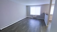 This single-floor space offers a clean, functional layout with a modern yet understated aesthetic. The main living area is finished with rich, dark hardwood-style flooring that runs throughout, providing a sharp contrast against the soft, neutral gray walls. The color palette creates a calm and cohesive atmosphere throughout the space.

Natural light enters through a large window, brightening the interior and contributing to an open, airy feel. A wall-mounted air conditioning unit is installed directly beneath the window, ensuring climate control during warmer months. Baseboard heating lines the perimeter of the room, offering efficient warmth during cooler seasons — a practical and unobtrusive heating solution.

The walls are smooth and freshly finished, with clean white baseboards framing the flooring and adding a crisp, polished edge to the overall look. The layout is open and versatile, allowing for a variety of furniture configurations to suit different needs.

Overall, the space presents a straightforward and well-maintained interior, combining practical climate features with contemporary finishes. The dark flooring, neutral tones, and ample natural light work together to create a bright yet grounded environment suitable for comfortable everyday living.