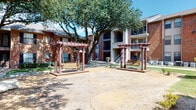 This multi-building residential complex showcases traditional brick construction across several two-story structures, creating a cohesive and well-established community aesthetic. The exteriors feature a combination of warm red brick and neutral siding accents, with private balconies and covered walkways accessible via staircases fitted with sturdy blue metal railings.

The grounds are a standout feature of the property, characterized by mature live oak trees that provide generous shade across the landscaped common areas. Neatly maintained lawns are complemented by decorative gravel borders and stone edging, creating a clean and organized outdoor environment. Paved concrete walkways connect the various buildings, guiding residents through the well-planned layout of the complex.

A central courtyard area serves as a gathering space, featuring two pergola-style wooden swing structures positioned on a stamped concrete pad — an inviting spot for relaxation. Picnic tables are conveniently placed beneath the tree canopy, offering shaded outdoor dining options. A gated pool area is also present, visible through black iron fencing and surrounded by additional pergola structures and lush greenery.

The overall ambiance of the property is one of a comfortable, established residential community, blending functional outdoor amenities with natural landscaping elements to create an inviting and livable environment.