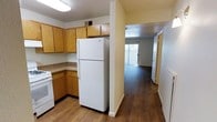 This two-floor residential space offers a practical and well-organized layout designed for comfortable everyday living. The kitchen is equipped with warm honey-toned wood cabinetry providing generous storage, a granite-style countertop, a white refrigerator with top freezer, and a white range, all set on durable hardwood-style flooring that flows seamlessly throughout the space. A structural column divides the kitchen from the adjacent hallway and living area, adding a sense of defined zones while maintaining an open feel. The hallway leads to the living area, which benefits from a sliding glass door or large window that invites natural light and connects the interior to the outdoors. The consistent hardwood-style flooring throughout enhances the cohesive and clean aesthetic of the space. Textured ceilings and neutral wall tones in a soft yellow-white palette contribute to a bright and inviting ambiance. The overall design is straightforward and functional, with a layout that prioritizes usability. Spanning two floors, the space provides ample room for both daily living and privacy, making it a well-rounded and informative example of practical residential design.