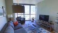 This single-floor space showcases a thoughtfully designed living area centered around dramatic floor-to-ceiling windows that frame expansive city skyline views, flooding the interior with abundant natural light. The living room is furnished with a plush grey sofa accented by vibrant coral throw pillows and a coordinating armchair, arranged around a sleek metal-framed coffee table. A geometric, multicolored area rug anchors the seating arrangement and adds a contemporary touch to the warm hardwood flooring beneath. Opposite the seating area, an open media console hosts a large flat-screen television, providing a functional entertainment focal point. A statement pendant lamp with an oversized drum shade adds a bold design element overhead. A small potted plant near the window introduces a natural accent against the urban backdrop visible through the glass. The walls are finished in a soft, neutral tone, complementing the modern aesthetic throughout. The overall ambiance is clean, bright, and urban-contemporary, making excellent use of its elevated position to highlight the dynamic cityscape beyond.