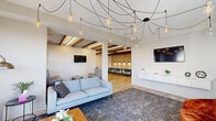 Located at 880 Maple Ave in Noblesville, Indiana, this thoughtfully designed single-floor amenity space blends modern aesthetics with practical functionality. The living room serves as the centerpiece, featuring a striking spider-style pendant chandelier with exposed Edison bulbs that drapes artfully across the ceiling. Large windows flood the space with natural light, highlighting the plush seating arrangements — including a light blue sofa, a rich burnt-orange accent chair, and a low-profile wood coffee table — all anchored by a textured area rug. A wall-mounted television and floating white media console add a polished, contemporary touch. An adjoining lounge area showcases warm wood beam ceilings and a light wood plank accent wall, with a long communal table and modern blue chairs ideal for collaborative seating. The kitchen features clean white subway tile backsplash, light wood cabinetry, and ample countertop workspace. The expansive exercise room is well-appointed with cardio machines, strength training equipment, free weights, resistance tools, and exercise balls, all set on durable carpet tile flooring with bright overhead lighting. A connecting hallway with tile flooring links the spaces seamlessly, completing this versatile and inviting facility.