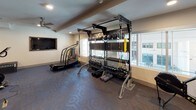 Located at 5619 Belmont Ave in Dallas, Texas, this well-appointed fitness center offers a comprehensive workout environment spread across a single floor. The space is thoughtfully organized into distinct functional zones, each designed to accommodate a wide range of exercise modalities.

The main workout area features a generous open floor plan covered in patterned, cushioned carpet, providing both comfort and durability underfoot. Overhead, ceiling fans and recessed lighting ensure proper ventilation and visibility throughout. A large framed wall mirror enhances the sense of space while allowing users to monitor their form during exercise.

The cardio section is equipped with multiple Peloton-style cycling bikes, a Woodway curved treadmill, and additional cardio machines, all positioned with ample spacing. Wall-mounted televisions are strategically placed for entertainment during workouts.

A dedicated strength training station includes a multi-functional RAX rack system stocked with kettlebells, dumbbells, medicine balls, resistance bands, and TRX suspension trainers, complemented by windows offering natural light and views of the surrounding Dallas streetscape.

A separate, glass-partitioned studio area provides space for group fitness classes or individual training, complete with stability balls, step platforms, and a cycling bike. The clean, neutral color palette and modern finishes create a focused, professional atmosphere throughout.