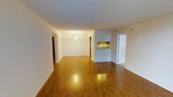 This single-floor space is characterized by an open-concept layout that promotes a sense of spaciousness and easy flow between living areas. Warm-toned hardwood flooring extends throughout, adding both visual continuity and a welcoming feel underfoot.

The main living and dining area is generously sized, with white walls and a clean, neutral palette that creates a bright and airy atmosphere. A pendant light fixture marks the dining area, providing focused illumination and a subtle decorative accent. The smooth, white ceiling complements the understated aesthetic of the space.

A notable architectural feature is the pass-through window connecting the kitchen to the main living area, offering both visual connectivity and practical functionality. A glimpse of the kitchen reveals stone or granite countertops, suggesting a well-appointed cooking space. Adjacent to this area, a hallway leads to additional rooms, including what appears to be a bathroom with glass-enclosed features. A second hallway extends toward further rooms, adding to the overall sense of depth and utility within this single-level layout. The overall ambiance is clean, functional, and move-in ready.