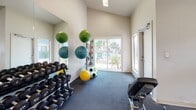 This fitness center is a comprehensively outfitted single-floor facility designed to support a wide range of workout routines. The space features soaring vaulted ceilings that lend an open, airy feel, complemented by natural light streaming in through multiple windows and a sliding glass door that offers views of an outdoor pool area and landscaping beyond.

The main workout area is divided into functional zones. The cardio section houses treadmills, an elliptical trainer, and a stationary bike, all positioned near wall-to-wall mirrors that visually expand the space and allow for form monitoring. A ceiling fan overhead aids in ventilation during intense sessions.

On the strength training side, a full dumbbell rack with a wide range of weights lines the mirrored wall, accompanied by kettlebells and a flat weight bench for versatile free-weight exercises. A multi-functional cable machine anchors a dedicated strength zone, offering pulley-based cable training for upper and lower body workouts.

Functional training is well supported with a collection of medicine balls in various sizes displayed on a wall-mounted rack, along with TRX suspension trainers and exercise mats. A wall-mounted mini-split air conditioning unit ensures climate control throughout.

Posted fitness center rules and informational signage reflect an organized, community-oriented facility. Dark rubber flooring throughout provides durability and safety, rounding out this practical and well-appointed fitness space.