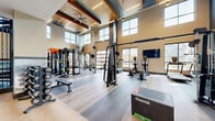 This expansive fitness center is designed with a contemporary aesthetic, characterized by soaring double-height ceilings, large multi-pane windows, and an open, airy layout that floods the space with natural light. Exposed wood-accented ceiling beams, recessed lighting, and ceiling fans contribute to a polished yet functional ambiance throughout.

The main workout floor features light wood-look luxury vinyl flooring and houses an extensive selection of Precor and other commercial-grade strength machines, including cable systems, a Smith machine, and a functional training rig equipped with TRX suspension trainers and resistance attachments. Plyo boxes, weight benches, and free weights are strategically distributed throughout the space.

A dedicated cardio zone along the windowed perimeter accommodates ellipticals, treadmills, and upright bikes, each positioned to offer outdoor views. Wall-mounted flat-screen televisions are placed throughout for entertainment.

A separate lower-level area features rubberized flooring and additional strength equipment, including a power rack and adjustable bench. Glass garage-style doors in one section open to an outdoor greenspace, seamlessly connecting interior and exterior training environments. An upper-level mezzanine with glass railings overlooks the main floor, adding architectural depth to the overall design.