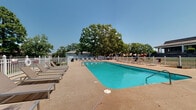 Located at 3807 McCain Park Dr in North Little Rock, Arkansas, this outdoor pool area offers a well-maintained recreational retreat. The centerpiece is a generously sized rectangular swimming pool with clear, vivid turquoise water, accessible via stainless steel entry rails at multiple points. The pool is surrounded by a broad concrete sun deck that provides ample space for relaxation and movement. Numerous lounge chairs and seating arrangements are distributed around the deck, offering guests comfortable spots to sunbathe or unwind. A white picket-style perimeter fence encloses the entire area, providing both security and a clean, classic aesthetic. Mature shade trees line the surroundings, offering natural canopy and a pleasant outdoor ambiance. A grill station is also available within the pool area, adding to its recreational functionality. The single-story building visible in the background features a low-pitched roof with wide overhangs, consistent with a hospitality or lodging property. The open-sky setting ensures abundant natural light throughout the day, making this pool area a bright and inviting outdoor space ideal for warm-weather enjoyment.