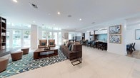 This contemporary single-floor amenity space is designed with an open-concept layout that seamlessly connects multiple functional zones, creating a bright and welcoming environment. The interior is finished with a sophisticated palette of white walls, light wood-look flooring, and large-format porcelain tile, lending the space a clean, modern aesthetic throughout.

The kitchen is a standout feature, anchored by an expansive granite-topped island with seating for several bar-height chairs upholstered in teal. Custom cabinetry combines white upper units with warm wood lower cabinets, complemented by a striking blue mosaic tile backsplash. Stainless steel appliances complete the well-appointed culinary area.

Adjacent to the kitchen, a spacious lounge area features comfortable sectional and accent seating arranged around a patterned navy-and-white area rug. A built-in wood bookshelf with integrated lighting adds warmth and a library-like quality to one corner. Large floor-to-ceiling sliding glass doors and clerestory windows flood the lounge with natural light and offer views of lush outdoor greenery.

A dedicated game room includes a billiards table and arcade machines, providing additional entertainment options. Linear pendant and recessed lighting throughout ensures even illumination, while curated artwork adds character and visual interest across the space.