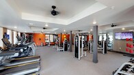 This fitness center in Little Elm offers a thoughtfully designed, single-floor workout facility built to accommodate a wide range of exercise routines. The expansive main floor is filled with an impressive array of strength training machines and cardio equipment, including treadmills, cable machines, and multi-function weight stations, all arranged to maximize usable floor space while maintaining open, comfortable circulation paths. The interior features a neutral-toned carpet floor and a tray ceiling accented with recessed lighting and multiple ceiling fans, ensuring both style and comfort during workouts. Bold orange accent walls add an energetic, motivating atmosphere to the space, while large mirrors along the walls enhance the sense of openness and allow for proper form checks. Structural columns are integrated seamlessly throughout the floor plan, lending an architectural rhythm to the layout. The combination of ample equipment variety, thoughtful lighting, and dynamic color choices makes this fitness center a well-rounded and inviting environment suited for both casual users and dedicated fitness enthusiasts.