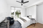 This thoughtfully designed single-floor studio offers a clean, contemporary aesthetic throughout. The main living and sleeping area is anchored by a built-in murphy bed that folds seamlessly into a white cabinetry wall, maximizing floor space and versatility. Light hardwood flooring runs throughout the unit, complementing the bright white walls and creating a warm, airy ambiance. A ceiling fan with a sleek matte black finish provides both style and function overhead. Large floor-to-ceiling windows flood the space with natural light and offer open views of the surrounding outdoor environment, enhancing the sense of openness. A cozy seating area with a navy blue accent chair and coordinating area rug adds a touch of color and comfort to the living space. The compact kitchenette is efficiently outfitted with modern gray cabinetry, dark granite-style countertops, a stainless steel refrigerator, built-in microwave, and an under-counter appliance — delivering a fully functional cooking space in a streamlined footprint. The bathroom features wood-look tile flooring, clean white walls, a wall-mounted sink, and a combined tub and shower area, all finished with matte black hardware accents for a cohesive, contemporary look throughout.