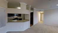This single-floor residence offers a thoughtfully designed open-concept layout that seamlessly connects the kitchen and living areas. The kitchen is a standout feature, showcasing dark espresso cabinetry paired with stainless steel appliances, including a microwave, electric range, and refrigerator. A curved breakfast bar with a neutral countertop serves as a natural divider between the kitchen and the main living space, providing both a functional prep area and an informal dining option. Crown molding runs throughout the space, adding a refined architectural touch to the ceilings and lending a sense of polish to the overall design. The living area is finished with plush, neutral-toned carpeting that contributes to a warm and comfortable ambiance. A hallway visible from the main area leads to additional rooms, suggesting a well-organized floor plan with clearly defined private and shared spaces. The consistent use of neutral wall tones throughout creates a cohesive and versatile backdrop, making the space feel open and inviting. Overall, this residence combines practical design elements with classic finishing details for a comfortable and functional living environment.