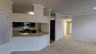 This single-floor residence offers a thoughtfully designed open-concept layout that seamlessly connects the kitchen and living areas. The kitchen is a standout feature, showcasing dark espresso cabinetry paired with stainless steel appliances, including a microwave, electric range, and refrigerator. A curved breakfast bar with a neutral countertop serves as a natural divider between the kitchen and the main living space, providing both a functional prep area and an informal dining option. Crown molding runs throughout the space, adding a refined architectural touch to the ceilings and lending a sense of polish to the overall design. The living area is finished with plush, neutral-toned carpeting that contributes to a warm and comfortable ambiance. A hallway visible from the main area leads to additional rooms, suggesting a well-organized floor plan with clearly defined private and shared spaces. The consistent use of neutral wall tones throughout creates a cohesive and versatile backdrop, making the space feel open and inviting. Overall, this residence combines practical design elements with classic finishing details for a comfortable and functional living environment.