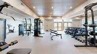 Located at 20-30 W Allens Lane in Philadelphia's Mt. Airy neighborhood, this fitness center offers a generously sized, single-floor exercise space designed with both functionality and aesthetic appeal in mind. The room is defined by its striking wood-paneled ceiling, which adds warmth and a modern architectural character to the otherwise clean, neutral interior. Recessed lighting throughout ensures the space is consistently well-lit, while large windows and glass doors along one wall invite ample natural light and provide views of the surrounding greenery.

The flooring consists of durable, speckled rubber-composite material suited for high-impact activity, and full-length mirrors along the walls enhance the sense of openness. The facility is comprehensively equipped for a wide range of workouts. Strength training options include TRX suspension systems, a power rack with bench, cable machines, a multi-station functional trainer, and a full rack of dumbbells. Cardio equipment includes treadmills, elliptical machines, and a stationary bike. An open floor area provides ample room for stretching or floor exercises. Restroom access is available directly within the facility. The overall ambiance is modern, clean, and inviting, making it a well-rounded fitness destination.
