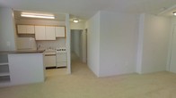 This single-floor unit presents an open and functional layout, with a clear visual connection between the living area and kitchen. The living space is finished with light-colored carpeting, creating a neutral and easy-to-maintain foundation. The kitchen is situated at one end of the space and is equipped with white cabinetry featuring wood-tone trim, a gas range, a sink, and under-counter appliances. Fluorescent lighting illuminates the kitchen workspace, while a flush-mount ceiling fixture provides ambient light to the adjoining hallway area. A partial wall or peninsula separates the kitchen from the carpeted living zone, maintaining an open feel while subtly defining the two areas. A hallway extending from the kitchen area leads to additional rooms, with a door visible at the far end, suggesting access to a bathroom or storage. The walls throughout are finished in a clean white, contributing to a bright, neutral interior. Overall, the space offers a simple, practical design with a cohesive flow between its main living and kitchen zones.