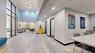 This single-floor amenity suite is thoughtfully designed to balance social engagement with focused productivity. The space is unified by a distinctive multi-toned plank flooring featuring gray, blue, and yellow accents that guides residents through each area seamlessly. Recessed lighting and drop ceilings maintain a clean, polished atmosphere throughout.

The main corridor opens into a welcoming lounge area anchored by a sectional sofa with mustard accent pillows, a terrazzo round coffee table, and a prominent circular branded wood wall installation. Bold, colorful pop-art prints are displayed throughout, adding dynamic visual energy to the neutral white walls.

Adjacent to the lounge, glass-walled rooms reveal a dedicated game room complete with a foosball table and pool table, as well as a social lounge with additional seating and a statement chandelier. A conference-style room features a long communal table with terrazzo detailing, surrounding chairs, a wet bar area, and textured accent wall.

Smaller private rooms offer wall-mounted flat-screen TVs and intimate seating configurations, ideal for quiet meetings or individual use. A compact meeting room with a round table, vivid yellow chairs, and a mounted display provides a bright, functional workspace. Bronze-framed glass doors throughout lend a refined, cohesive architectural detail to the entire suite.