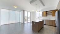 This contemporary single-floor space showcases a clean, open-concept layout that seamlessly integrates the kitchen and living areas. Large-format grey porcelain tiles span the entire floor, contributing to a cohesive and polished aesthetic throughout. The kitchen is outfitted with warm wood-toned cabinetry that contrasts elegantly against crisp white countertops, while a central island provides additional workspace and serves as a natural dividing point between the kitchen and living zones. Pendant lighting fixtures hang above the island, adding focused illumination and a touch of modern style.

The living area is defined by expansive floor-to-ceiling sliding glass doors and frosted panel windows that flood the space with abundant natural light. These doors open onto a private balcony, offering panoramic views of the surrounding cityscape and greenery beyond. The white ceiling features a subtle recessed detail above the kitchen area, adding architectural interest. The overall ambiance is bright, airy, and minimalist, making it a versatile space suited for both everyday living and entertaining.