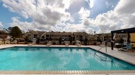 Located in Robertsdale, Alabama, this outdoor community pool area offers a well-appointed recreational space designed for relaxation and leisure. The centerpiece is a generously sized swimming pool with clear, blue water, framed by a broad, light-colored concrete sundeck that provides ample space for sunbathing and socializing. Multiple lounge chairs are arranged around the pool, offering comfortable spots to unwind while taking in views of the surrounding residential community and open sky above.

A covered pavilion with white columns anchors one side of the deck, providing shaded seating and a protected gathering area, complete with patio furniture for casual outdoor dining. A water vending machine is conveniently accessible near the pavilion entrance, adding a practical touch for pool-goers. A pergola-style shade structure with privacy curtains is also present, offering an additional retreat from the sun.

The pool area is enclosed by a black metal fence, with manicured landscaping and young trees lining the perimeter, lending the space a clean and inviting character. Access to the adjacent clubhouse or pool facility building is provided through elegant, multi-pane French doors with keypad entry, ensuring both security and style. The overall ambiance is bright, open, and community-oriented, making it an ideal outdoor amenity for residents of the Robertsdale community.
