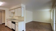 This single-floor residence offers a clean, open-concept layout that seamlessly connects the kitchen and living areas, creating a cohesive and practical living environment. The kitchen is outfitted with white cabinetry that extends to the upper walls, providing ample storage space. A light-toned countertop runs along the workspace, which includes a stainless steel sink and a black dishwasher tucked neatly beneath. The kitchen is partially separated from the main living area by a half-wall partition, adding a subtle sense of definition while maintaining an open, airy feel between the two spaces. The living area is generously proportioned, with neutral white walls that offer a versatile backdrop for a variety of interior styles. Rich, dark hardwood-style flooring flows continuously throughout both the kitchen and living spaces, adding warmth and visual continuity. The smooth white ceilings and clean wall surfaces keep the interior bright and welcoming. Electrical outlets are thoughtfully positioned along the walls, offering flexibility for furniture arrangement. Overall, the space presents a straightforward, well-maintained interior that balances functionality with an inviting, comfortable atmosphere suitable for everyday living.