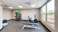 Located at 4805 W Genesee St in Syracuse, NY, this fitness center offers a focused, single-room exercise space designed to meet a range of workout needs. The room features a drop tile ceiling with recessed panel lighting that keeps the space bright and functional. Durable speckled flooring covers the entire area, providing a practical surface suited for both equipment use and floor exercises. Large windows along one wall flood the space with natural light and offer open views of the surrounding streetscape, creating an airy atmosphere despite the room's compact footprint. The fitness center is equipped with a treadmill, elliptical trainer, and recumbent stationary bike for cardio training, alongside a dedicated weight bench for strength work. A well-organized equipment rack holds an assortment of dumbbells, medicine balls, and a stability ball, providing versatile options for functional fitness routines. Neutral-toned textured walls contribute to a clean, modern aesthetic throughout. A windowed door provides access to an adjoining area, and a small storage cabinet near the entrance keeps informational materials and essentials organized. Overall, this fitness center delivers a practical, well-lit, and thoughtfully equipped space for residents or tenants seeking convenient on-site exercise facilities.