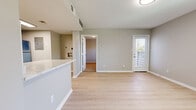 This single-floor residence offers a thoughtfully designed layout that balances open communal spaces with comfortable private quarters. The living area is finished with light hardwood-style flooring and neutral greige walls, creating a clean, contemporary atmosphere. A exterior door with slatted blinds allows natural light to fill the space while providing privacy. The open-concept design connects the living area seamlessly to the kitchen, which is anchored by a peninsula with a light stone-style countertop. The kitchen is well-equipped with gray shaker-style cabinetry, a white subway tile backsplash, and a full suite of stainless steel appliances including a refrigerator, range, and over-the-range microwave. A skylight-style overhead fixture provides bright, even illumination throughout the cooking space. The bedrooms are carpeted in a warm neutral tone, offering a soft contrast to the hard flooring in the main living areas. One bedroom benefits from a pair of windows that invite natural light and outdoor views, while the primary bedroom includes access to a private en-suite bathroom featuring gray vanity cabinetry. The unit is finished with clean white trim and baseboards throughout, lending a polished, cohesive look to the entire space.