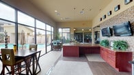 This single-floor community lounge area is designed to serve as a welcoming and functional gathering space. The interior is anchored by a prominent granite-topped counter finished in rich mahogany cabinetry, providing ample workspace and storage. A striking natural limestone column serves as a key architectural focal point, complemented by a decorative mosaic tile accent wall that houses wall-mounted flat-screen televisions. Additional cabinetry with granite countertops and a tile backsplash lines one wall, offering a convenient refreshment station stocked with various amenities. Warm yellow walls and high ceilings contribute to an open, airy atmosphere, while recessed and pendant lighting provide balanced illumination throughout. Large floor-to-ceiling windows and glass doors flood the interior with natural light and offer expansive views of the outdoor pool area, complete with lounge chairs and a spa. Bar-height seating near the windows allows residents to enjoy the poolside scenery from within. A secondary entry with rustic wood-framed doors provides direct access to the pool deck, where a generously sized swimming pool and surrounding patio with lounge seating await. The space seamlessly connects indoor comfort with outdoor recreation.