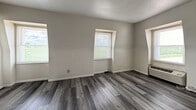 Located at 4025 US-45 S in Mattoon, IL, this single-floor studio unit offers 320 square feet of functional living space. The living area features dark gray laminate wood flooring, neutral gray walls, a textured ceiling, and two windows fitted with blinds that allow natural light to fill the room. A wall-unit HVAC system provides climate control, and built-in white shelving along one wall offers convenient storage and display space.

The kitchen area transitions to tile flooring and is equipped with essential appliances, including a white refrigerator, a white electric range, and a sink set into wooden cabinetry with dark countertops. A flush-mount ceiling light provides overhead illumination, and the layout flows openly into the living space, maximizing the utility of the compact floor plan.

The bathroom features full-wall square tile in a neutral gray tone, a standard white toilet, and a tub-shower combination with a white surround. A towel bar adds practical functionality to the space.

Overall, this unit presents a straightforward and efficiently arranged layout suited for single occupancy, with durable flooring, included kitchen appliances, and ample natural light throughout the living area.