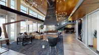 The Riverwalk lobby is a bold architectural statement that blends industrial character with refined, contemporary design. Upon entry, a branded welcome mat sets the tone beneath a dramatic entryway flanked by floor-to-ceiling glass doors that flood the space with natural light and frame urban skyline views beyond.

The heart of the lobby is anchored by a commanding central fireplace with a large, sculptural metal hood that draws the eye upward toward soaring ceilings clad in warm wood paneling. A series of sputnik-style chandeliers punctuate the vertical space, casting an inviting ambient glow throughout. Exposed industrial ductwork runs overhead, adding a raw, loft-inspired contrast to the refined finishes below.

The lounge area features curated seating—plush sofas, leather accent chairs, and a live-edge coffee table—arranged atop a richly patterned area rug. Large windows wrap the perimeter, offering panoramic city views and abundant daylight. A glass-railed staircase with wooden treads leads to an upper mezzanine level, beneath which a cozy dining nook with woven chairs and a Sputnik pendant creates an intimate gathering space. Backlit onyx wall panels and textured stone surfaces add layers of warmth and sophistication throughout this impressively appointed communal space.