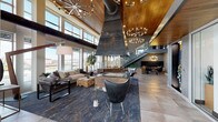 The Riverwalk lobby is a bold architectural statement that blends industrial character with refined, contemporary design. Upon entry, a branded welcome mat sets the tone beneath a dramatic entryway flanked by floor-to-ceiling glass doors that flood the space with natural light and frame urban skyline views beyond.

The heart of the lobby is anchored by a commanding central fireplace with a large, sculptural metal hood that draws the eye upward toward soaring ceilings clad in warm wood paneling. A series of sputnik-style chandeliers punctuate the vertical space, casting an inviting ambient glow throughout. Exposed industrial ductwork runs overhead, adding a raw, loft-inspired contrast to the refined finishes below.

The lounge area features curated seating—plush sofas, leather accent chairs, and a live-edge coffee table—arranged atop a richly patterned area rug. Large windows wrap the perimeter, offering panoramic city views and abundant daylight. A glass-railed staircase with wooden treads leads to an upper mezzanine level, beneath which a cozy dining nook with woven chairs and a Sputnik pendant creates an intimate gathering space. Backlit onyx wall panels and textured stone surfaces add layers of warmth and sophistication throughout this impressively appointed communal space.