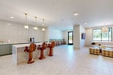 Located at 2700 N Lake Forest Dr in McKinney, TX, this thoughtfully designed single-floor community lounge space offers a refined and welcoming atmosphere through a cohesive blend of modern and organic design elements. The open layout is anchored by large-format hexagonal tile flooring in a soft cream tone that flows seamlessly throughout the entire space.

The kitchenette area serves as a functional centerpiece, featuring sage green cabinetry with gold hardware, a white quartz countertop island, full-height subway tile backsplash, stainless steel appliances, and a dedicated beverage station. A trio of gold pendant lights with frosted globe shades hang above the island, illuminating the sculptural hand-carved wooden bar stools that serve as a striking artistic focal point.

The lounge area transitions naturally into two distinct seating zones: one anchored by a curved cream sectional sofa paired with a round wood coffee table beneath an elegant gold chandelier, and another featuring a long caramel-toned banquette flanked by modern white accent tables. Brass wall sconces and framed artwork add warmth and personality to the walls.

Natural light filters in through black-framed windows and a glass entry door, offering views of the surrounding greenery. A wall-mounted flat-screen TV provides an additional entertainment feature. The overall ambiance is sophisticated yet approachable, making this a versatile and inviting communal gathering space.