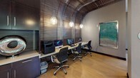 Located at 2009 N 14th Street in Arlington, Virginia, this single-floor space is a thoughtfully designed amenity suite that blends functionality with striking contemporary aesthetics. The business center is a standout feature, showcasing an architecturally dramatic arched ceiling clad in horizontal wood slat paneling that curves elegantly overhead. A built-in workstation accommodates multiple computer workstations side by side, illuminated by a series of sculptural glass pendant lights that cast a warm, focused glow across the light hardwood flooring. Dark cabinetry and storage solutions line the walls, providing a polished, professional atmosphere. Floor-to-ceiling glass partitions and exterior-facing windows flood the space with abundant natural light, offering views of the landscaped outdoor courtyard with mature trees and curated plantings. A cozy lounge nook features a round table flanked by upholstered chairs beneath another decorative wood slat panel, creating an intimate setting for informal meetings or quiet moments. Original artwork in cool, oceanic tones adorns the walls, adding a curated artistic touch throughout. Signage identifying the space as "Fusion" is prominently displayed near the entrance. The overall ambiance is modern and refined, combining warm wood tones, sleek metallic accents, and carefully selected furnishings for an inviting yet professional environment.
