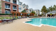 This expansive outdoor pool amenity in West Columbia, South Carolina offers a well-appointed retreat designed for relaxation and recreation. The centerpiece is a generously sized swimming pool with crystal-clear turquoise water, featuring a smooth paver deck that surrounds the pool on all sides, providing ample space for lounging and socializing.

A striking wooden pergola lines one side of the pool deck, providing shaded relief and framing a long row of sling-back lounge chairs — ideal for sunbathing or unwinding. Teal market umbrellas are stationed throughout the deck, offering additional shade at various seating clusters. Additional lounge seating is arranged along the opposite side of the pool, ensuring plenty of accommodation for guests.

A distinct lower patio area features a stacked-stone accent wall with a built-in water feature, brick raised planters, and bistro-style high-top tables and chairs, creating a more intimate gathering space. Lush tropical plantings, including palm trees and manicured shrubs, add vibrant greenery throughout the area.

The pool area is enclosed by an ornamental iron fence and is surrounded by multi-story residential buildings with classic brick and neutral-toned exteriors, balconies, and pitched rooflines. The overall ambiance is polished, welcoming, and resort-inspired, making it a standout communal outdoor amenity.