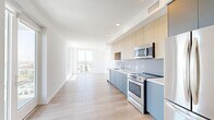 Located on S Western Ave in the heart of Los Angeles, this modern one-bedroom, one-bathroom unit offers a thoughtfully designed open-concept layout finished with light wood-style flooring throughout. The combined kitchen and living area forms the heart of the unit, where a sleek, contemporary kitchen stands out with two-tone cabinetry—pale wood uppers paired with powder-blue lower cabinets—complemented by a glossy tile backsplash, white quartz countertops, and a full suite of stainless steel appliances including a range, over-the-range microwave, dishwasher, and refrigerator. The open living area extends toward expansive floor-to-ceiling windows that frame sweeping views of the Los Angeles cityscape, flooding the space with abundant natural light. The well-proportioned bedroom continues the bright, airy aesthetic with large windows offering additional city views. A hallway connects the living spaces to the bathroom, which features large-format tile flooring, full-wall tile surrounding a soaking tub and shower, and clean, modern fixtures. A dedicated in-unit laundry closet with a stacked washer and dryer adds everyday convenience. The overall design is clean, minimalist, and modern, creating a comfortable and functional urban living environment.