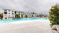 Located at 275 Pine Terrace Dr in Conroe, TX, this outdoor amenity space is designed as a resort-style retreat within a modern residential community. The centerpiece is a generously sized freeform swimming pool with vivid turquoise water, gentle entry steps, and a wading area, all framed by a wide, light-toned concrete pool deck. Rows of sleek lounge chairs line the pool's perimeter, complemented by blue market umbrellas that provide shade throughout the day.

The surrounding recreational areas offer a variety of outdoor living options. A dedicated lounge zone features stylish hanging egg chairs arranged around a small fire table, creating an intimate gathering spot. A canopied daybed cabana provides a shaded retreat for relaxation overlooking the pool and landscaped grounds. Additional sun loungers are scattered across open deck areas with views of the multi-story white-painted residential buildings that enclose the courtyard.

A covered outdoor dining terrace runs along the building's façade, equipped with ceiling fans and large communal dining tables backed by floor-to-ceiling dark-framed windows. Manicured landscaping with ornamental grasses and young trees adds greenery throughout the space. The overall aesthetic is clean and contemporary, combining white board-and-batten architectural details with dark iron fencing for a polished, cohesive outdoor environment.
