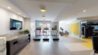 This fitness center is designed to accommodate a comprehensive workout experience across two distinct zones. The cardio area features wood-look flooring and houses multiple treadmills with individual screens, stationary bikes equipped with interactive Expresso displays, and elliptical trainers. Recessed lighting is complemented by decorative yellow-accented ceiling fixtures, while a large wall-mounted television and built-in cabinetry provide added convenience. Glass doors at the end of the cardio zone open directly to an outdoor pool and lounge area, allowing natural light to flood the space.

The strength training zone transitions to a bold, geometric flooring pattern in yellow, grey, and tan, anchored by a striking metallic column. Floor-to-ceiling mirrors line the walls, visually expanding the space. A full dumbbell rack, weight benches, functional training equipment, and cable machines are thoughtfully arranged throughout. A textured yellow accent wall adds a design focal point to one end of the room. The overall aesthetic is clean and contemporary, with neutral walls and bright, even lighting that creates an energetic yet focused environment suitable for varied fitness routines.