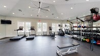 This fitness center offers a comprehensive workout environment designed to accommodate a wide range of exercise needs. The space features vaulted ceilings accented by a large statement ceiling fan, recessed lighting, and multiple windows with French doors that flood the interior with natural light and provide views of the surrounding outdoor greenery. The durable rubber flooring spans the entire facility, providing a safe and functional surface for all types of training.

The cardio zone is outfitted with multiple treadmills, stair climbers, and an elliptical machine, all positioned to take advantage of the natural light from the windows. Full-length wall mirrors visually expand the space while assisting users with form and technique. A dedicated strength training area houses a multi-station cable machine and an adjustable weight bench, complemented by a mounted flat-screen television. The free weight section features an extensive rack of dumbbells in a range of sizes, kettlebells, and an assortment of medicine and stability balls. TRX suspension training straps are also available, adding versatility to functional fitness routines. An open floor area with exercise mats provides ample space for stretching, yoga, or bodyweight workouts. The overall ambiance is clean, modern, and welcoming.