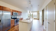 This efficiently designed one-bedroom, one-bathroom unit offers a thoughtful layout across a single floor. The open-concept kitchen and living area serves as the heart of the space, featuring rich hardwood flooring throughout. The kitchen is outfitted with warm-toned wood cabinetry, a mosaic tile backsplash, stainless steel appliances including a refrigerator, range, and built-in microwave, and a generous granite-topped island with a stainless steel sink and pendant lighting above — ideal for both meal prep and casual dining. The living area benefits from expansive floor-to-ceiling windows that flood the space with natural light and offer sweeping urban views of the surrounding cityscape. A glass-panel door leads to an outdoor access point, further enhancing the connection to the exterior. The bedroom is carpeted for comfort and includes a window with additional urban views, a dedicated closet, and direct access to the bathroom. The bathroom features clean, light-toned tile flooring and a glass-enclosed walk-in shower. A utility room provides added convenience. Track lighting and pendant fixtures contribute to the modern, urban aesthetic found throughout the unit.