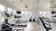 Located in Glendale, Arizona, this fitness center offers a clean, modern exercise environment designed to support a well-rounded workout routine. The space features light wood-look flooring, crisp white walls, and floor-to-ceiling mirrors that visually expand the room and allow users to monitor their form during exercise. Dual ceiling fans circulate air throughout, contributing to a comfortable workout atmosphere.

Large windows line multiple walls, flooding the space with abundant natural light and providing views of the surrounding outdoor landscape, including palm trees and the community grounds. A wall-mounted flat-screen television is positioned for easy viewing from the cardio equipment area.

The fitness center is equipped with a comprehensive selection of exercise machines, including multiple treadmills, elliptical trainers, stationary and recumbent bikes, and additional cardio equipment. A dedicated free weights area features a dumbbell rack alongside an adjustable weight bench, supporting strength training needs.

Glass doors on one end of the room open directly to an outdoor courtyard area, enhancing the connection to the outdoors and providing easy access to exterior amenities. The overall design of the space is bright, functional, and well-organized, offering residents a practical and motivating environment for daily fitness activities.