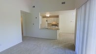 This two-story residence is designed with an open and airy floor plan that immediately draws attention to its notably high vaulted ceilings, which lend the main living area a spacious, expansive feel. The living space is finished with wall-to-wall carpeting in a soft, neutral tone, creating a comfortable and cohesive atmosphere throughout.

At the heart of the main level sits a well-appointed kitchen, thoughtfully positioned behind a breakfast bar-style pass-through counter that connects it directly to the living area. The kitchen features white cabinetry extending to the upper walls, light countertops, and white appliances including a range, providing a clean and functional workspace. Overhead lighting in the kitchen ensures the area remains well-lit. A pendant light fixture is also visible, adding a subtle decorative touch near the dining area.

The open sightlines between the kitchen and living spaces promote a sense of connectivity, making the layout well-suited for everyday living and entertaining. Neutral wall colors throughout complement the light finishes and contribute to the overall bright, welcoming ambiance of the home.