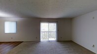 This single-floor space offers a straightforward and functional layout designed for comfortable living. The main living area is finished with wall-to-wall carpeting in a neutral tone, providing a soft and welcoming underfoot feel that complements the room's understated color palette of soft whites and warm grays.

A standout feature of the living area is the sliding glass door, which serves as both a source of abundant natural light and a direct access point to an outdoor balcony. The view through the door reveals lush, verdant greenery, creating a seamless connection between the interior and the natural surroundings outside. Vertical blinds frame the door, offering adjustable light control and privacy.

The walls are smooth and neutral, providing a clean backdrop that suits a variety of interior styles. The ceiling features a subtle textured finish, adding a touch of dimension to the otherwise minimalist overhead surface. The overall ambiance of the space is calm and bright, with natural light playing a key role in enhancing the open feel of the room. The simple, uncluttered design makes it an adaptable space, well-suited for a range of furnishing arrangements and personal aesthetics.