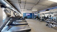 Located at 360 N First St in Minneapolis, MN, this fitness center offers a comprehensive workout facility spread across a single, open floor. The space is thoughtfully divided into distinct workout zones, each designed to support a variety of fitness routines.

The cardio zone lines one side of the room and is outfitted with multiple treadmills and elliptical machines, all positioned beside large windows that invite natural light and provide views to the outside. A wall-mounted flat-screen TV keeps exercisers entertained during their sessions.

The strength training area occupies the opposite side and features a robust selection of equipment, including power racks, cable machines, a variety of weight benches, and a dumbbell rack. Full-length mirrors along the walls enhance the sense of space and allow for proper form checks during workouts.

A dedicated spin and functional training alcove is set apart from the main floor, furnished with stationary cycling bikes and a weight bench, set against a striking blue accent wall adorned with a decorative relief design. Rubber flooring in this area provides durability and safety.

Throughout the space, exposed ductwork and industrial-style fluorescent lighting contribute to a clean, modern aesthetic. The bold blue accent walls and neutral carpet flooring create a cohesive, motivating atmosphere well-suited for daily fitness activity.