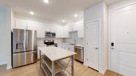 This well-appointed single-floor unit offers a clean, modern aesthetic defined by bright white walls, light hardwood flooring, and recessed lighting throughout. The open-concept layout seamlessly connects the kitchen and living area, creating a spacious and airy feel. The kitchen is a standout feature, outfitted with crisp white shaker-style cabinetry, quartz countertops, a classic white subway tile backsplash, and a full suite of stainless steel appliances including a French door refrigerator with ice and water dispenser, electric range, over-the-range microwave, and dishwasher. A freestanding kitchen island with open shelving provides additional prep and storage space. The living area is generously sized, illuminated by natural light from dual windows and complemented by a ceiling fan with integrated lighting. The bathroom features white shaker-style cabinetry with brushed gold hardware, a quartz vanity top with an undermount sink, a large framed mirror, and a glass-enclosed walk-in shower. A dedicated laundry area houses a full-size top-loading washer and dryer. Additional storage is provided by a spacious walk-in closet fitted with wire shelving. The overall ambiance is clean, bright, and move-in ready.