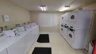 This single-floor shared laundry facility is designed for practicality and efficiency. A row of top-loading washing machines lines one wall, each equipped with its own faucet connections, providing ample capacity for multiple users at once. Opposite the washers, a bank of stacked dryer units maximizes the available floor space, offering several drying options simultaneously.

The room features light-toned tile flooring that is easy to maintain, with black floor mats placed at key entry points to manage tracked-in debris. The walls are finished in a neutral beige tone, contributing to a clean and functional atmosphere. Overhead, fluorescent lighting fixtures provide bright, even illumination throughout the space.

A window fitted with white vertical blinds is centered on the far wall, allowing natural light to supplement the artificial lighting while maintaining privacy. The overall layout is straightforward and well-organized, with clear circulation space between the machines for comfortable use. The facility is clearly intended to serve multiple users, with the arrangement and quantity of machines reflecting a communal-use design.
