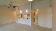 This single-floor residence features an open-concept layout that connects the living area to the kitchen through a practical pass-through design. The living space is finished with wall-to-wall carpeting in a neutral beige tone, contributing to a clean and cohesive aesthetic throughout.

The kitchen is partially enclosed by a half-wall partition, which also serves as a breakfast bar equipped with a granite-style countertop and two corbel-supported shelves — ideal for casual dining or additional counter space. The kitchen itself is outfitted with warm wood-toned cabinetry, a white refrigerator, and a tile backsplash, providing a functional and well-organized cooking area.

Wall-mounted sconce lighting within the kitchen nook adds a warm glow, complementing the ceiling-mounted light fixtures visible in the adjoining spaces. A ceiling fan is installed in the living area, offering both air circulation and ambient light.

A hallway visible beyond the kitchen leads to additional areas of the unit, suggesting a practical and efficient floor plan. Overall, the space presents a neutral, move-in-ready environment with straightforward finishes suited for everyday living.