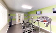 Located at 4827 Jersey Pike in Chattanooga, TN, this fitness center is a well-equipped, single-floor facility designed to support a well-rounded cardio workout routine. The space is defined by its vibrant aesthetic, featuring bold horizontal green and gray stripes along the walls that create an energizing atmosphere. Overhead, flush-mounted fluorescent lighting ensures the room is brightly and evenly lit throughout.

The facility is lined with a comprehensive selection of cardio machines, including multiple treadmills, elliptical trainers, and a recumbent bike, all arranged in a row to maximize the use of the elongated floor plan. A large wall mirror at one end visually expands the space and allows users to monitor their form during workouts. Multiple wall-mounted flat-screen televisions are positioned throughout for entertainment during exercise sessions.

Natural light filters in through windows fitted with blinds along one wall, adding a welcoming brightness to the interior. The room is accessed through elegant French doors with glass panes, providing a clean and polished entryway. Dark flooring anchors the space and complements the lively wall colors. A water cooler station is also conveniently available. The textured ceiling and clean white trim complete the well-maintained, functional design of this community fitness center.