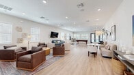 Located on Halo Bend Court in Wesley Chapel, Florida, this single-story community clubhouse offers a generously sized, open-plan living space designed for both relaxation and recreation. The interior is finished with light hardwood-style flooring throughout, crisp white walls, and recessed lighting that creates a bright, welcoming atmosphere.

The main living area is thoughtfully arranged into multiple distinct seating zones, each furnished with a curated mix of sofas, armchairs, and coffee tables in neutral and complementary tones. A wall-mounted flat-screen TV serves as a focal point for one of the lounge areas, while in-ceiling speakers add to the entertainment capabilities of the space.

Toward the rear of the room, a full-sized billiards table takes center stage alongside high-top seating, a beverage refrigerator, and a tiled accent wall, creating a dedicated game and entertainment zone. Large windows fitted with white blinds line the walls, allowing ample natural light to filter throughout the space.

A set of glass-panel double doors provides direct access to an outdoor area, offering views of what appears to be a pool or courtyard beyond. The entry foyer features matching double doors with grid-pane windows, shiplap wall detailing, and a console table with decorative accents, setting an inviting tone upon arrival. Overall, the space strikes a balance between casual comfort and community functionality.
